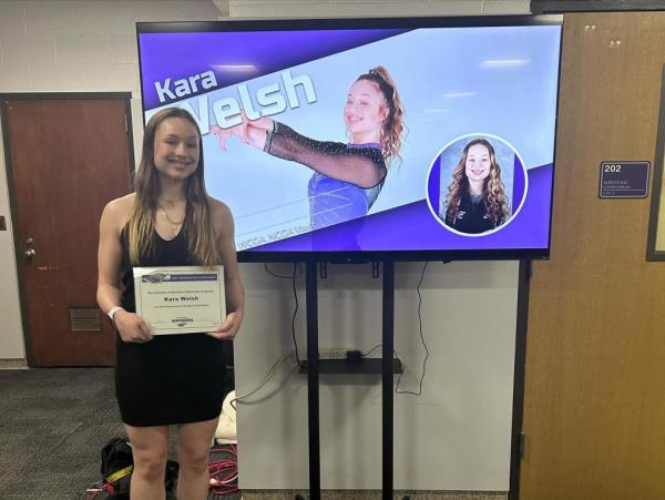 Kara Welsh holds a certificate in front of a TV screen showing her name and photo